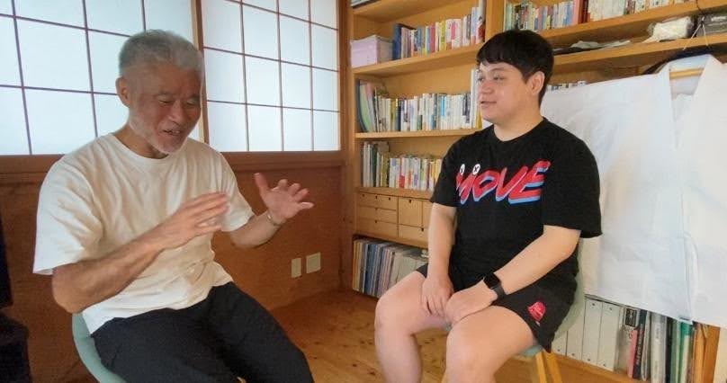 本棚のある室内で、年配の男性と若い男性が向かい合って対話している様子。