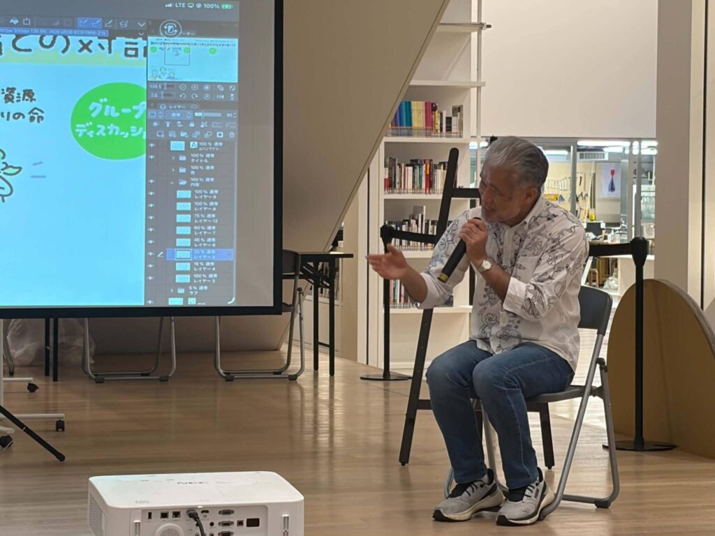 スクリーンと本棚のある会場で、白髪の男性がマイクを持って話している様子。